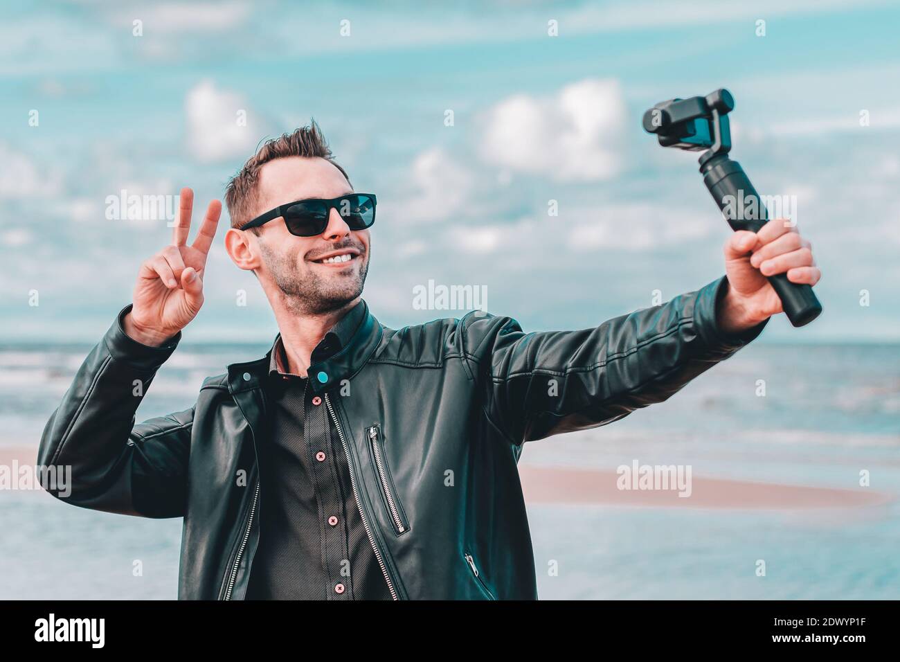 Youthful Blogger in occhiali da sole che fanno Selfie o video in streaming  alla spiaggia usando Action Camera con stabilizzatore di fotocamera gimbal.  Bell'uomo in vestiti neri fare foto contro il mare