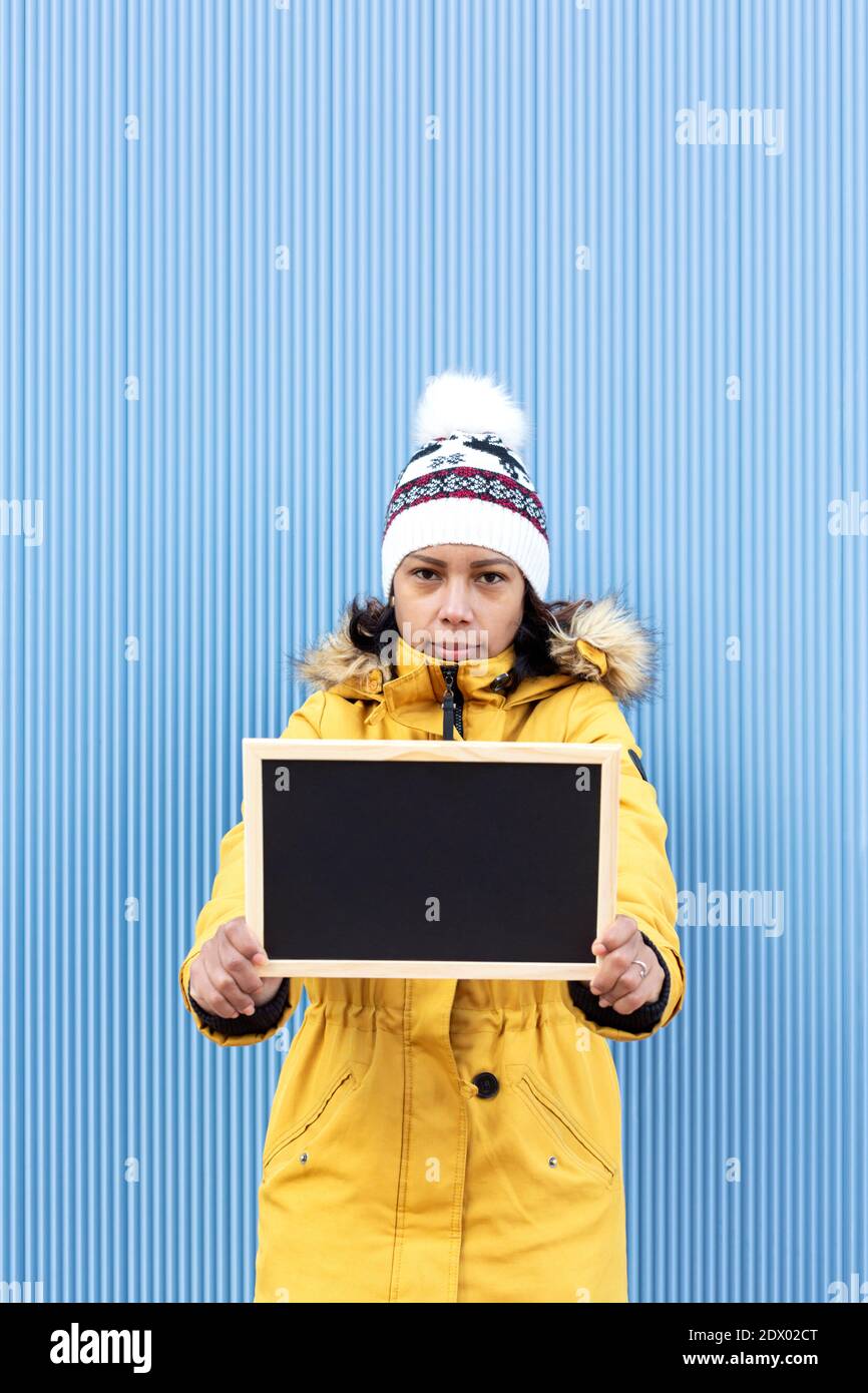 Ritratto verticale di una donna latina seria in abiti invernali con una lavagna bianca. È in piedi accanto a un muro blu. Spazio per il testo. Foto Stock