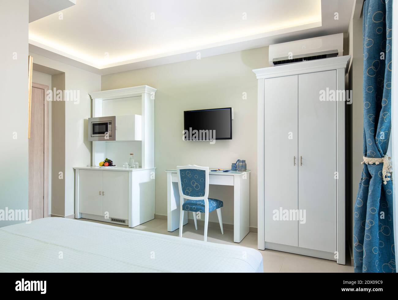 Interni in stile classico bianco e blu della camera d'hotel con armadio a modulo cucina, armadio e tavolo TV in un unico spazio di un piccolo studio moderno Foto Stock
