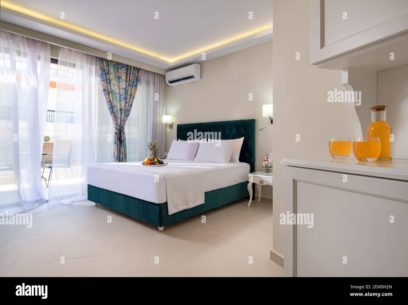 Elegante e moderno interno della camera da letto nell'appartamento dell'hotel a doppio suit con morbido letto in tessuto color acquamarina, mobili in stile classico bianco e fruttato di benvenuto Foto Stock