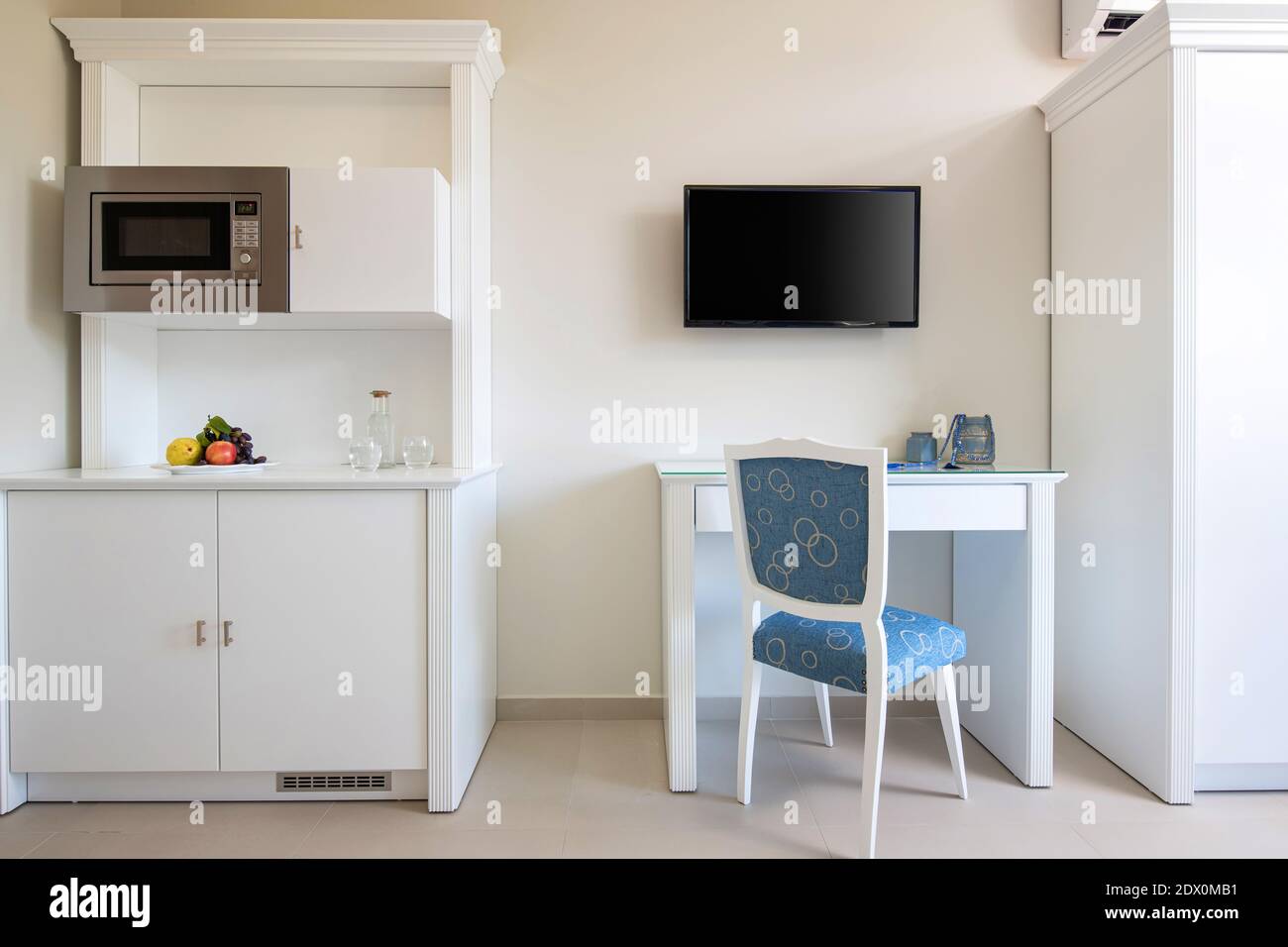 Vista frontale della parete bianca con TV, tavolo, sedia classica blu, armadio cucina in legno con elettrodomestici in piccola stanza interni moderni Foto Stock