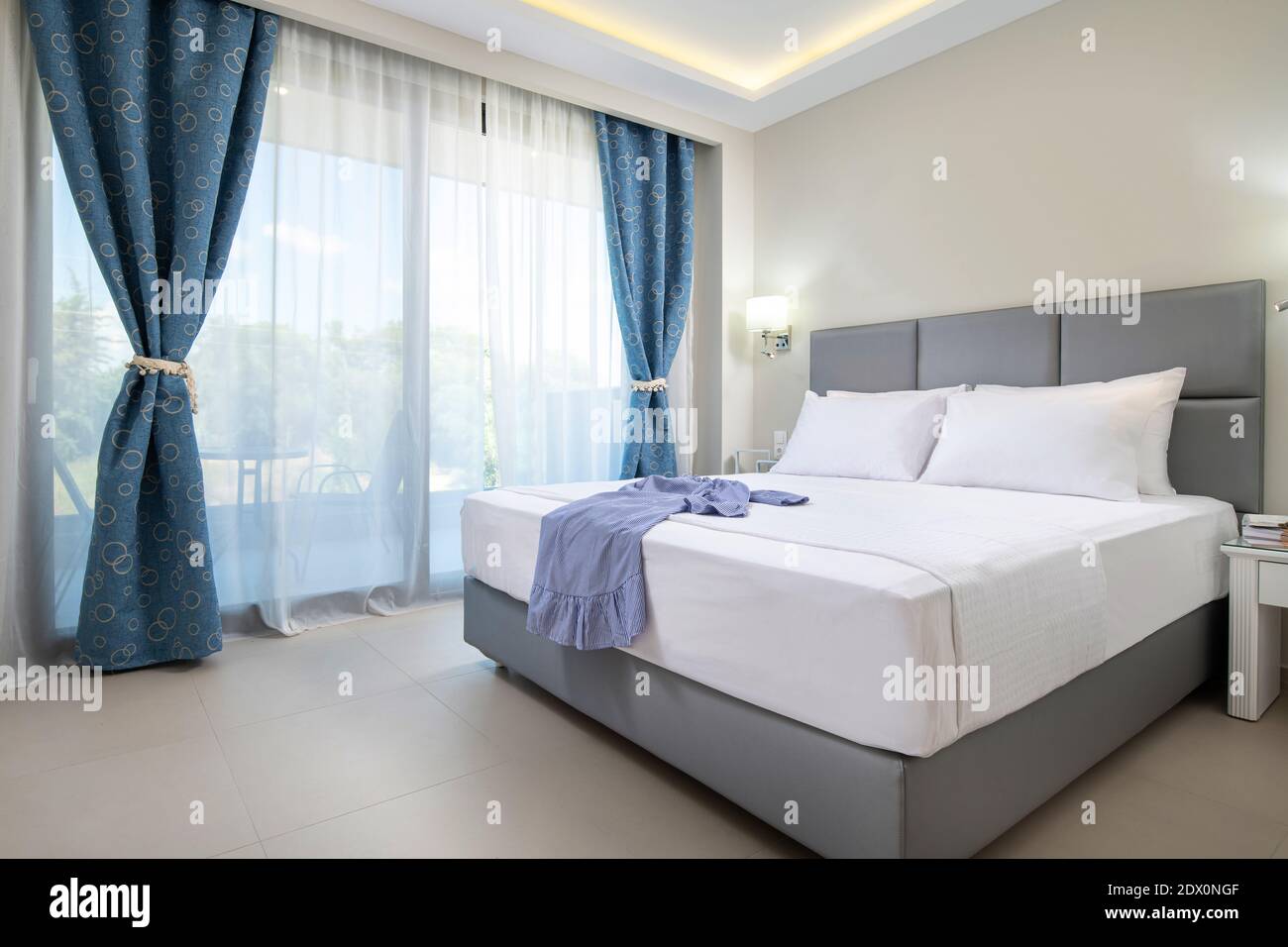 Interni dal design moderno di eleganti camere da letto con letto matrimoniale in tessuto grigio chiaro, biancheria da letto bianca e tende blu in stile classico appartamento dell'hotel Foto Stock