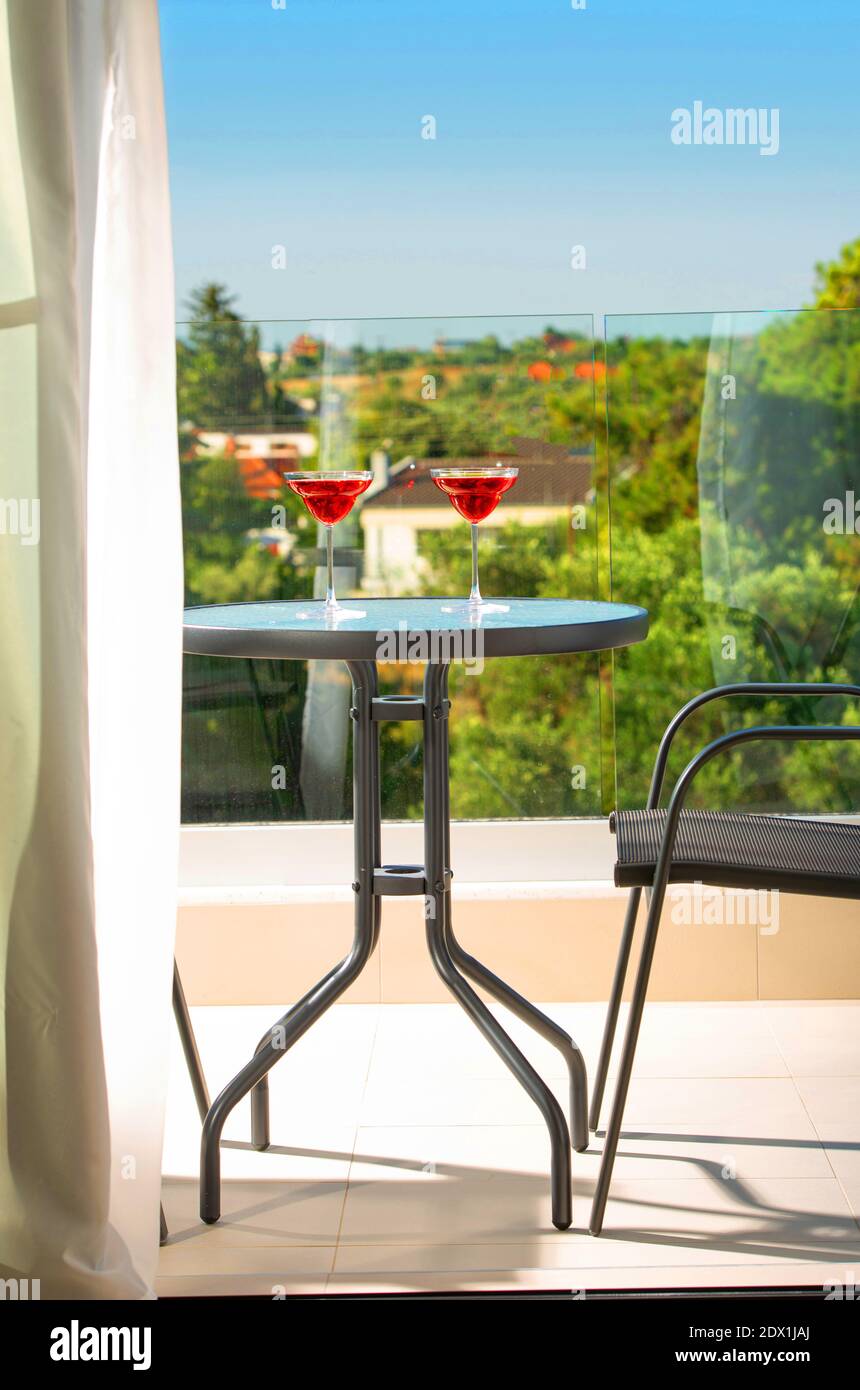 Primo piano di balcone aperto con vista sul giardino estivo. Terrazza patio con tavolo in vetro, sedie e vetri rossi margarita Foto Stock