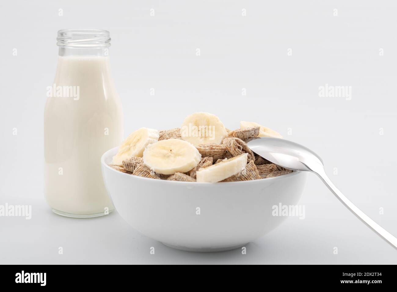 Colazione sana e ricca fonte di calcio e fibra concetto con cereali generici di grano trinciato e fette di banana, bottiglia di latte e cucchiaio isolato Foto Stock