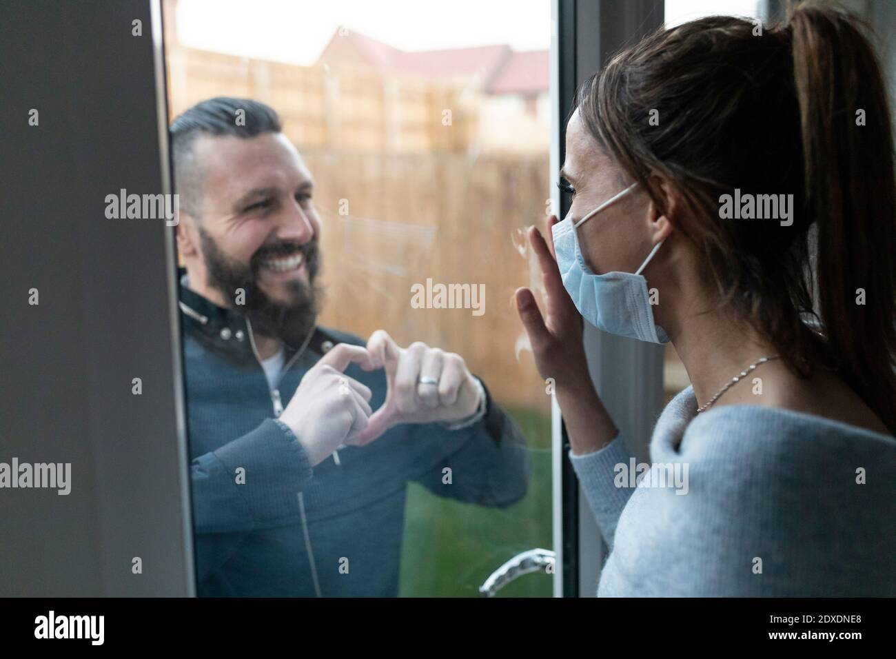 Il ragazzo mostra il movimento della forma del cuore alla fidanzata attraverso il vetro della finestra Durante la crisi COVID-19 Foto Stock