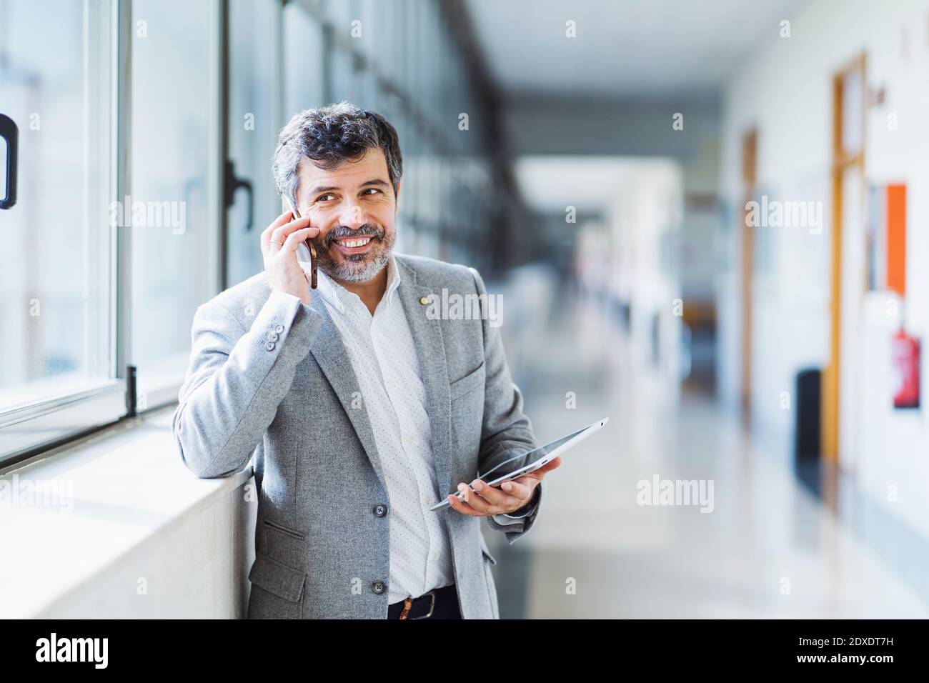 Sorridente professore guardando via mentre risponde telefono in corridoio a. università Foto Stock