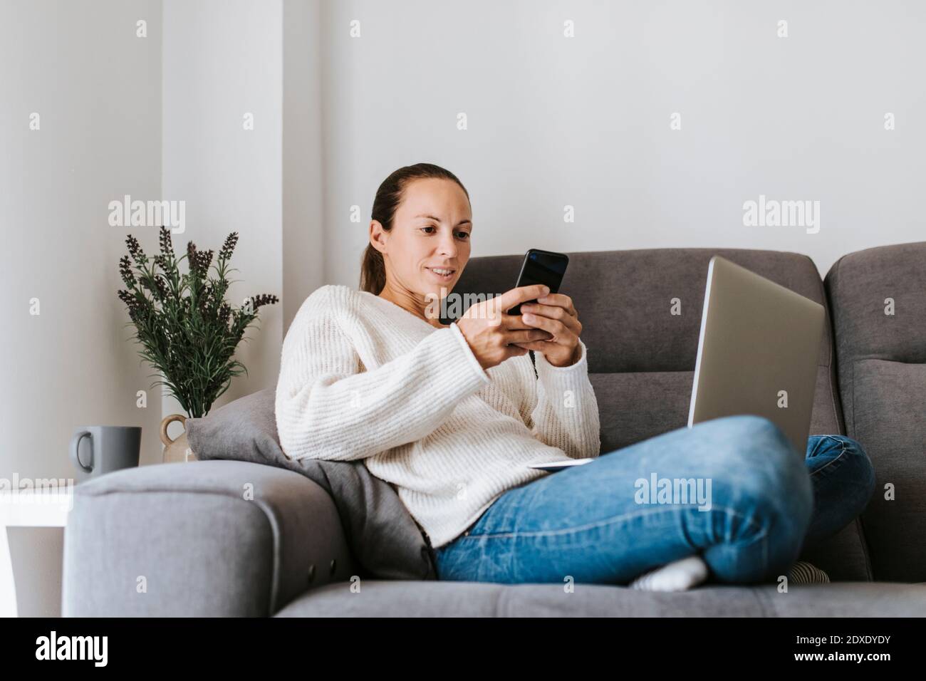 Messaggi di testo per donna tramite smartphone mentre si è seduti con il computer portatile sul divano Foto Stock
