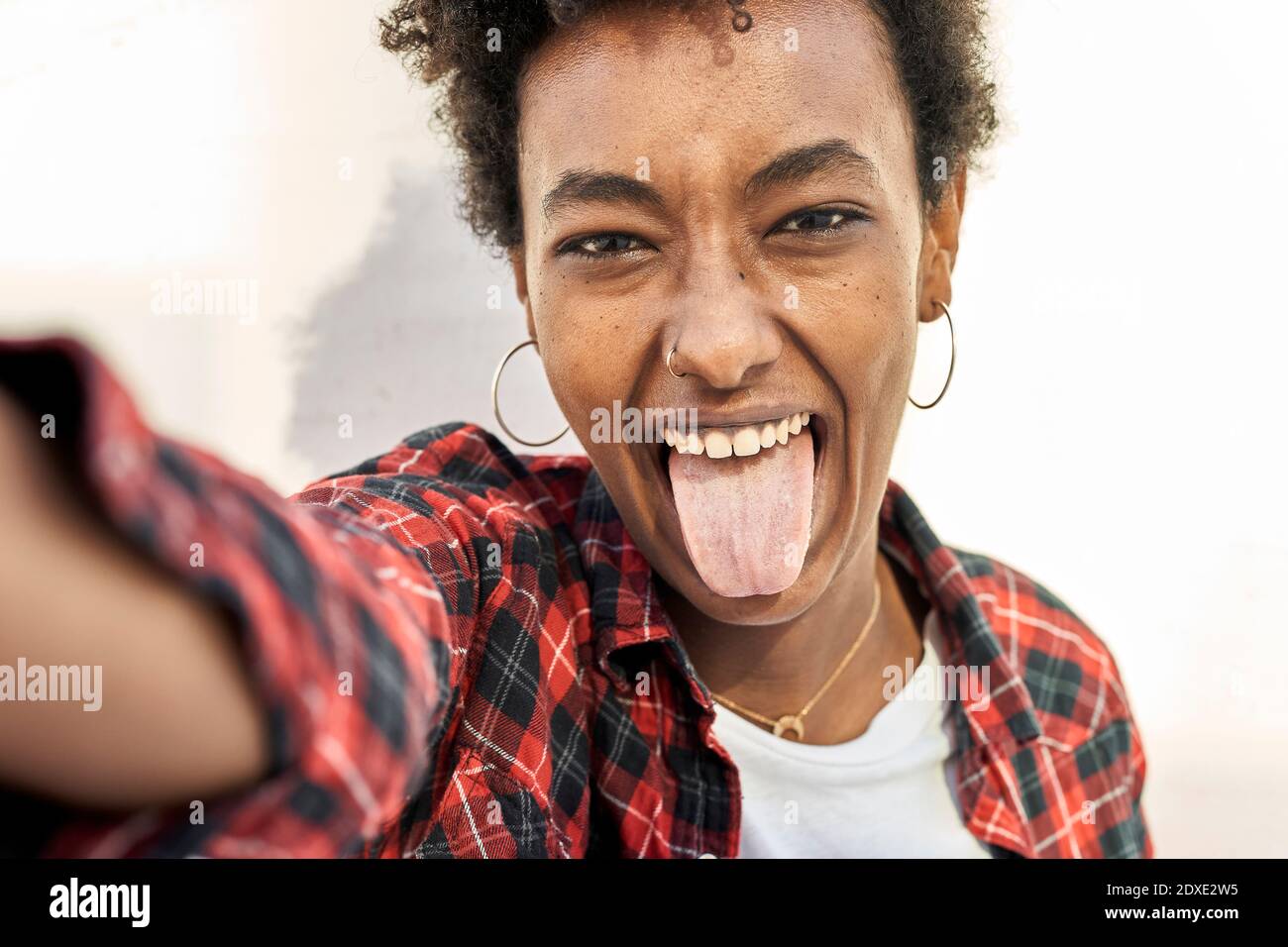 Giovane donna allegra che prende selfie mentre si attacca la lingua contro parete bianca Foto Stock