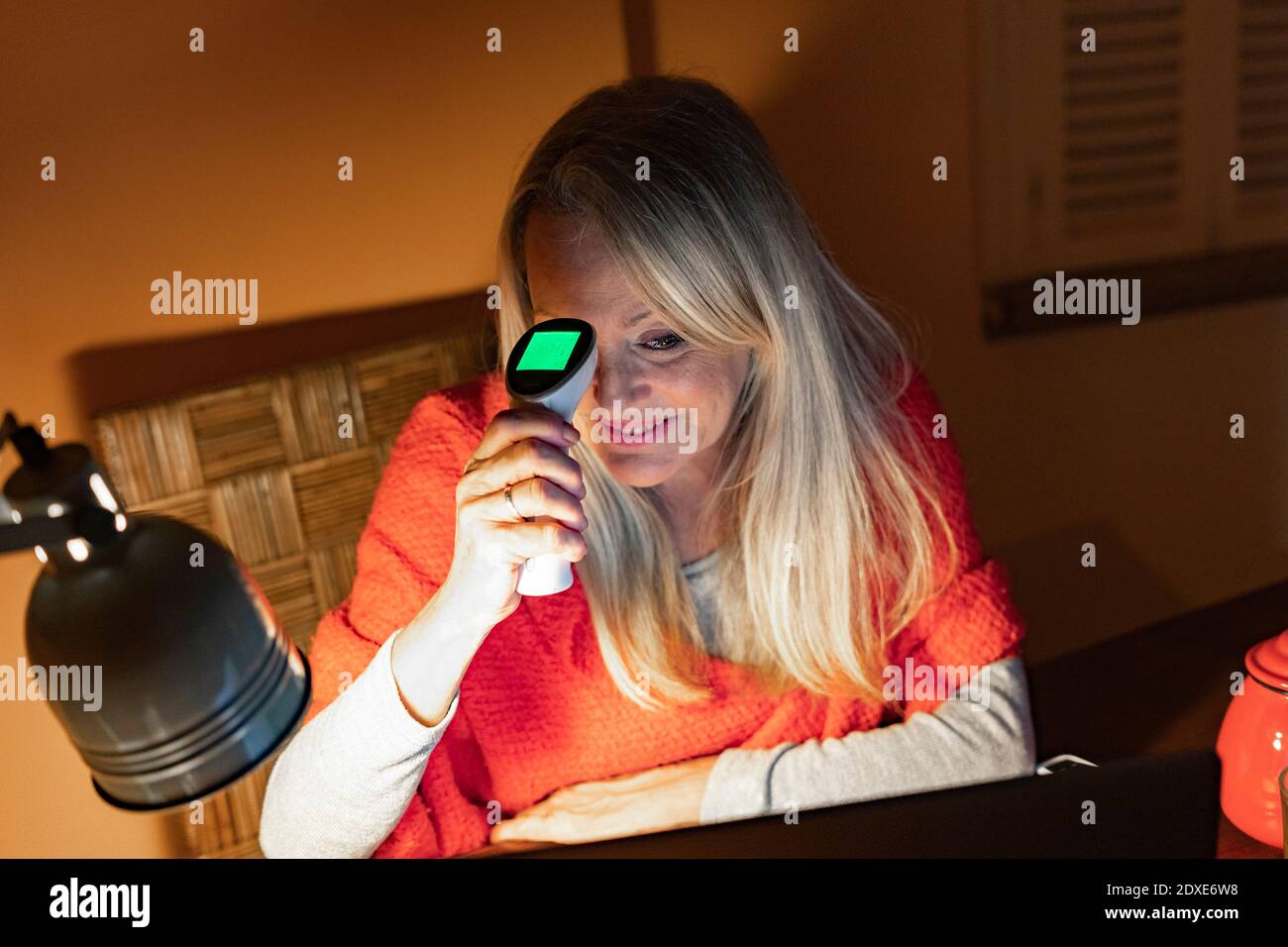 Donna anziana sorridente che usa un termometro a infrarossi mentre guarda il video chiama il computer portatile a casa Foto Stock