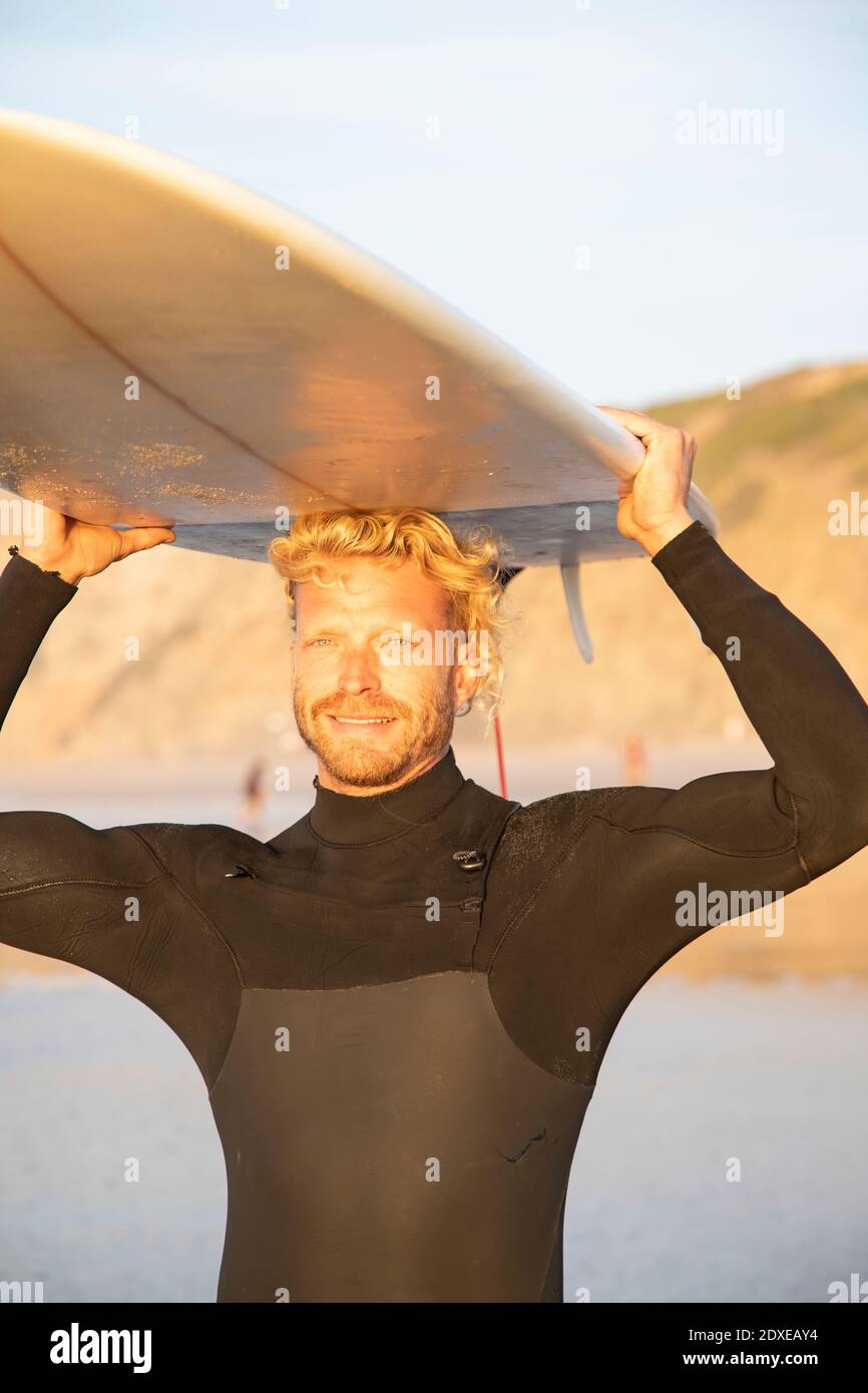 Uomo biondo sorridente che porta la tavola da surf sopra la testa alla spiaggia durante tramonto Foto Stock