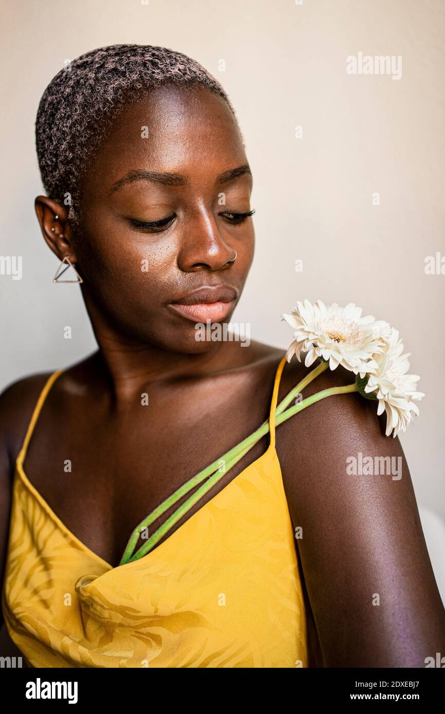 Hipster femminile con capelli corti sbiancati guardando fiore sopra spalla contro parete bianca Foto Stock