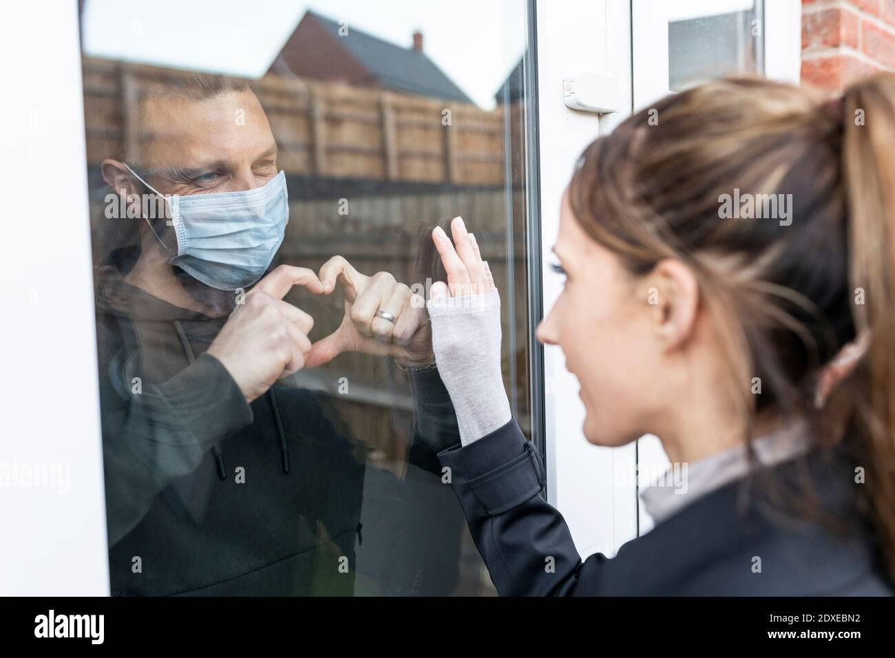 Il ragazzo mostra il movimento della forma del cuore alla fidanzata attraverso il vetro della finestra mentre messo in quarantena a casa Foto Stock