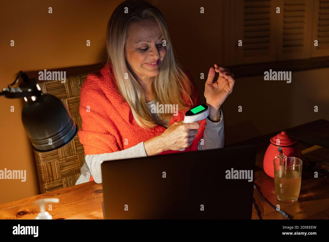 Donna sorridente che controlla la temperatura con un termometro a infrarossi mentre si è seduti con computer portatile a casa Foto Stock