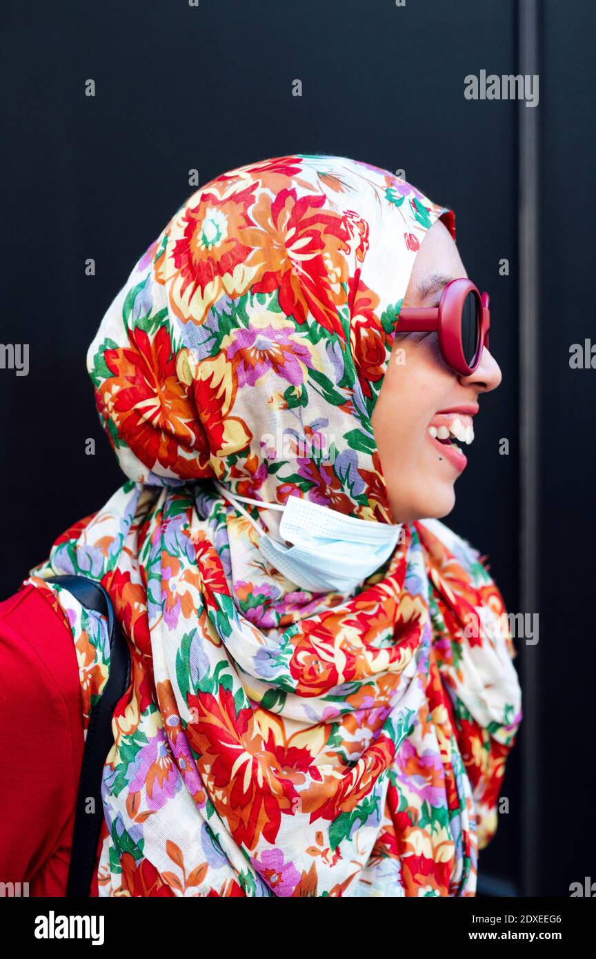 Donna allegra in hijab floreale contro parete nera durante COVID-19 Foto Stock