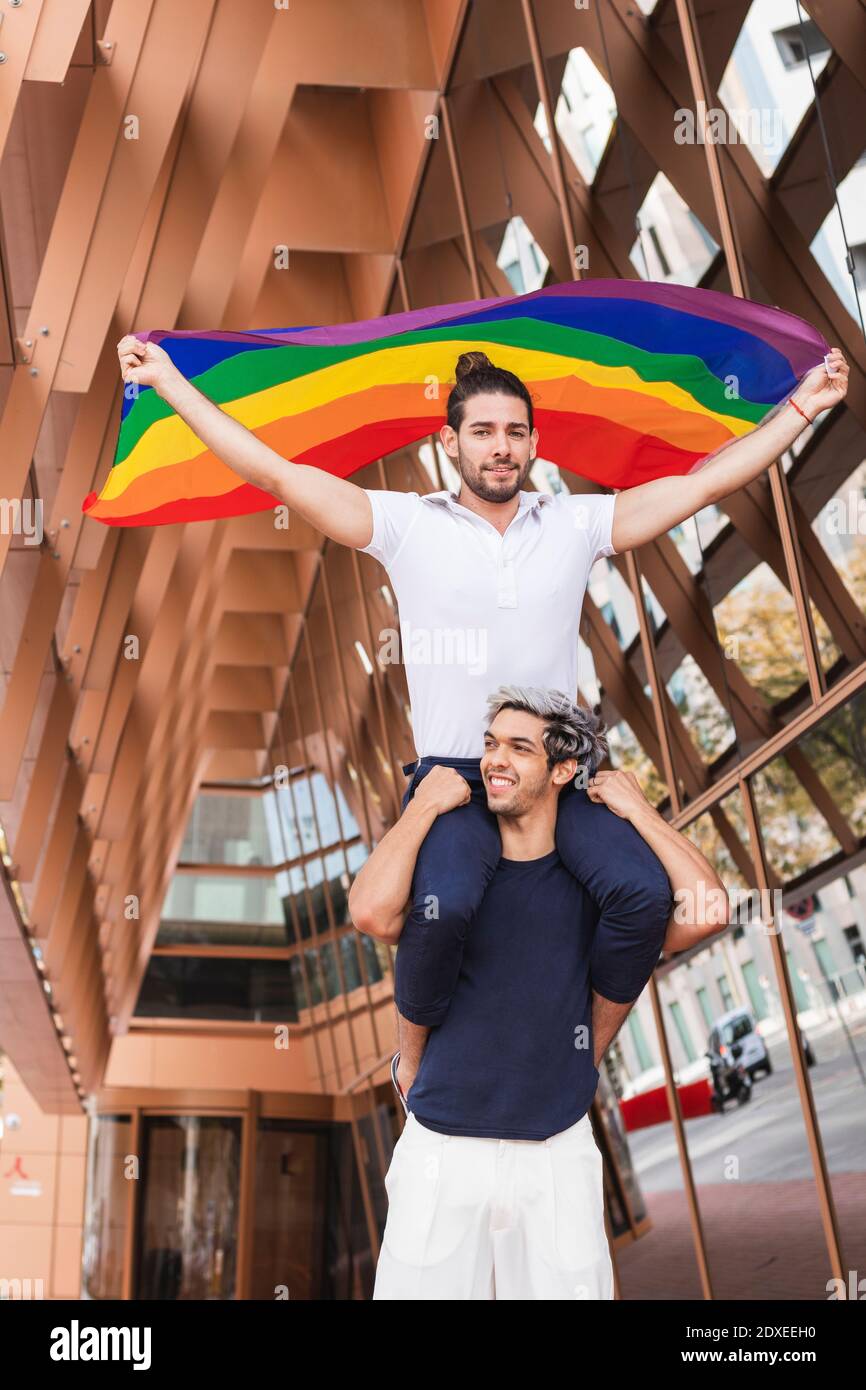 Uomo sorridente che porta il compagno di sesso maschile con sciarpa  arcobaleno sulle spalle in città Foto stock - Alamy