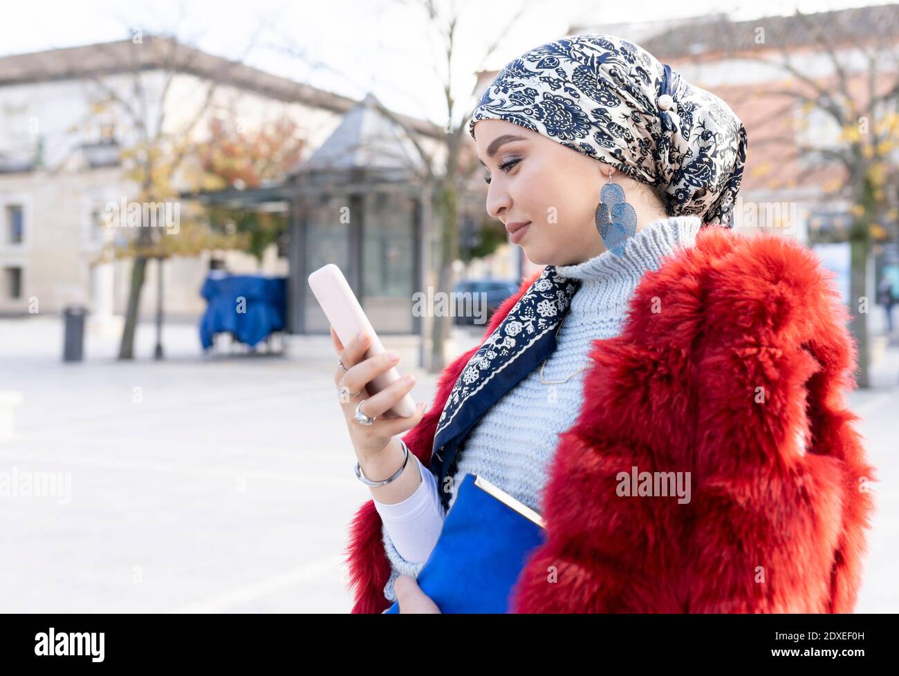 Donna sorridente che indossa foulard e giacca in pelliccia utilizzando il telefono cellulare mentre si è in città Foto Stock