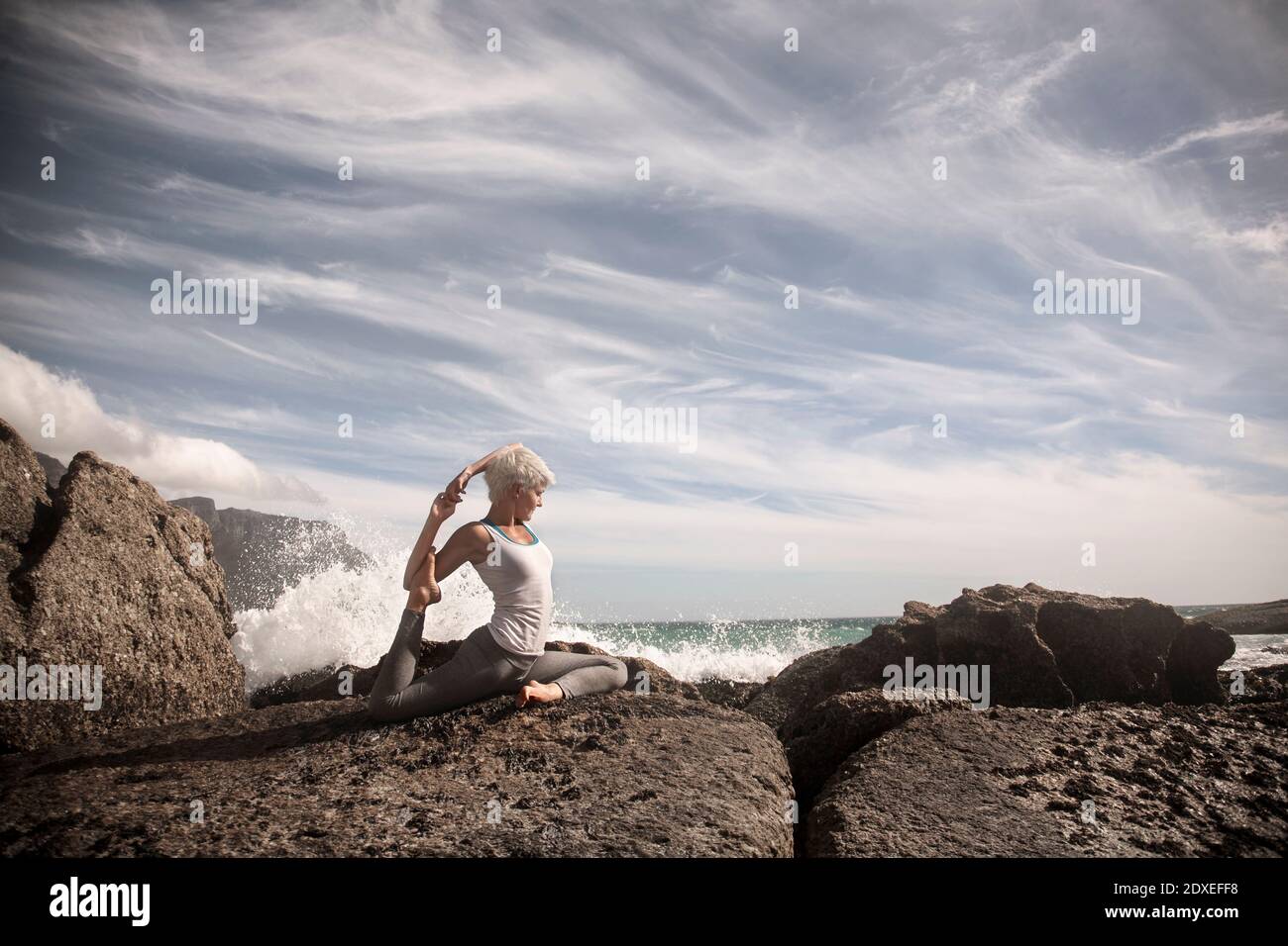 Donna bionda flessibile che pratica yoga sulla formazione rocciosa in spiaggia Foto Stock