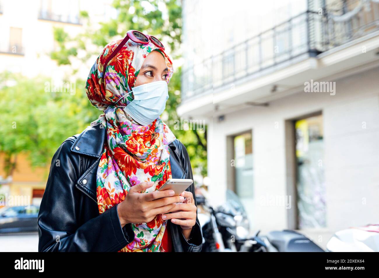 Donna in hijab floreale che tiene il telefono cellulare guardando via dentro città durante il coronavirus Foto Stock