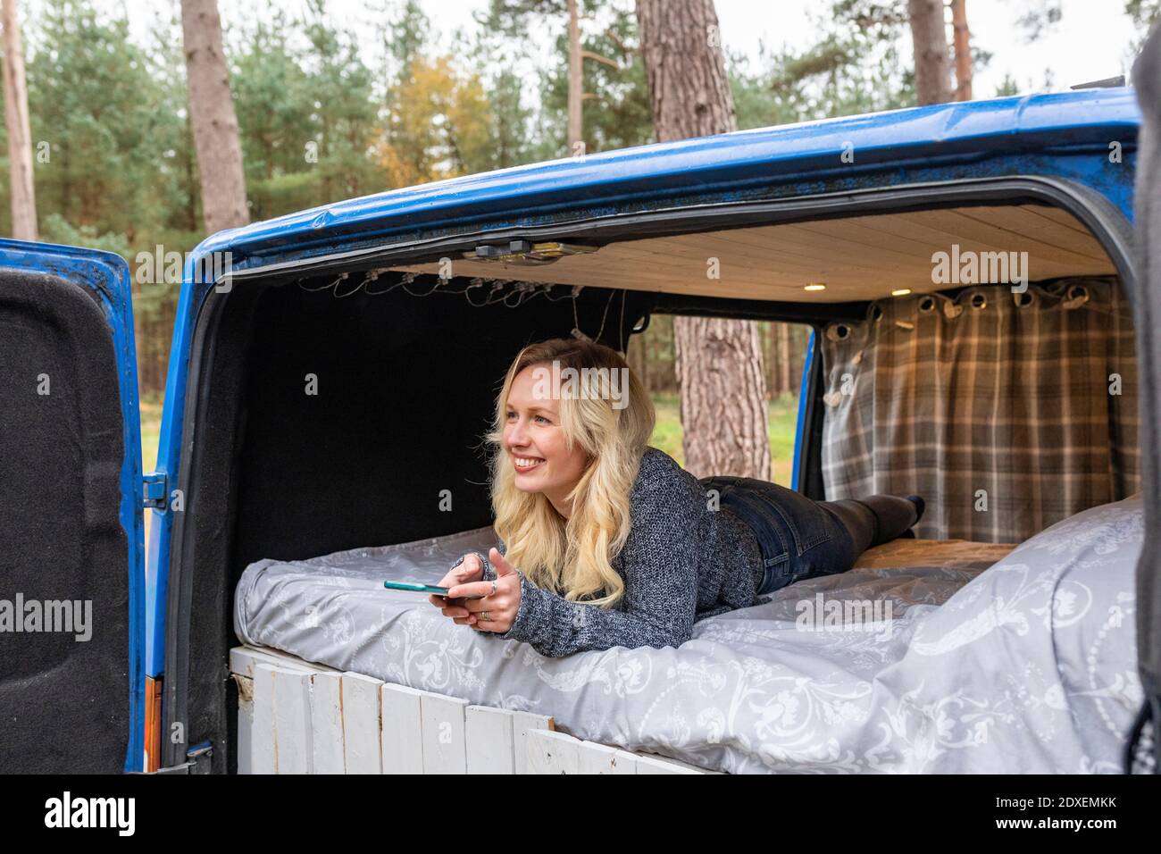 Donna felice con telefono cellulare giorno sognare mentre si sdraiava letto in furgone Foto Stock