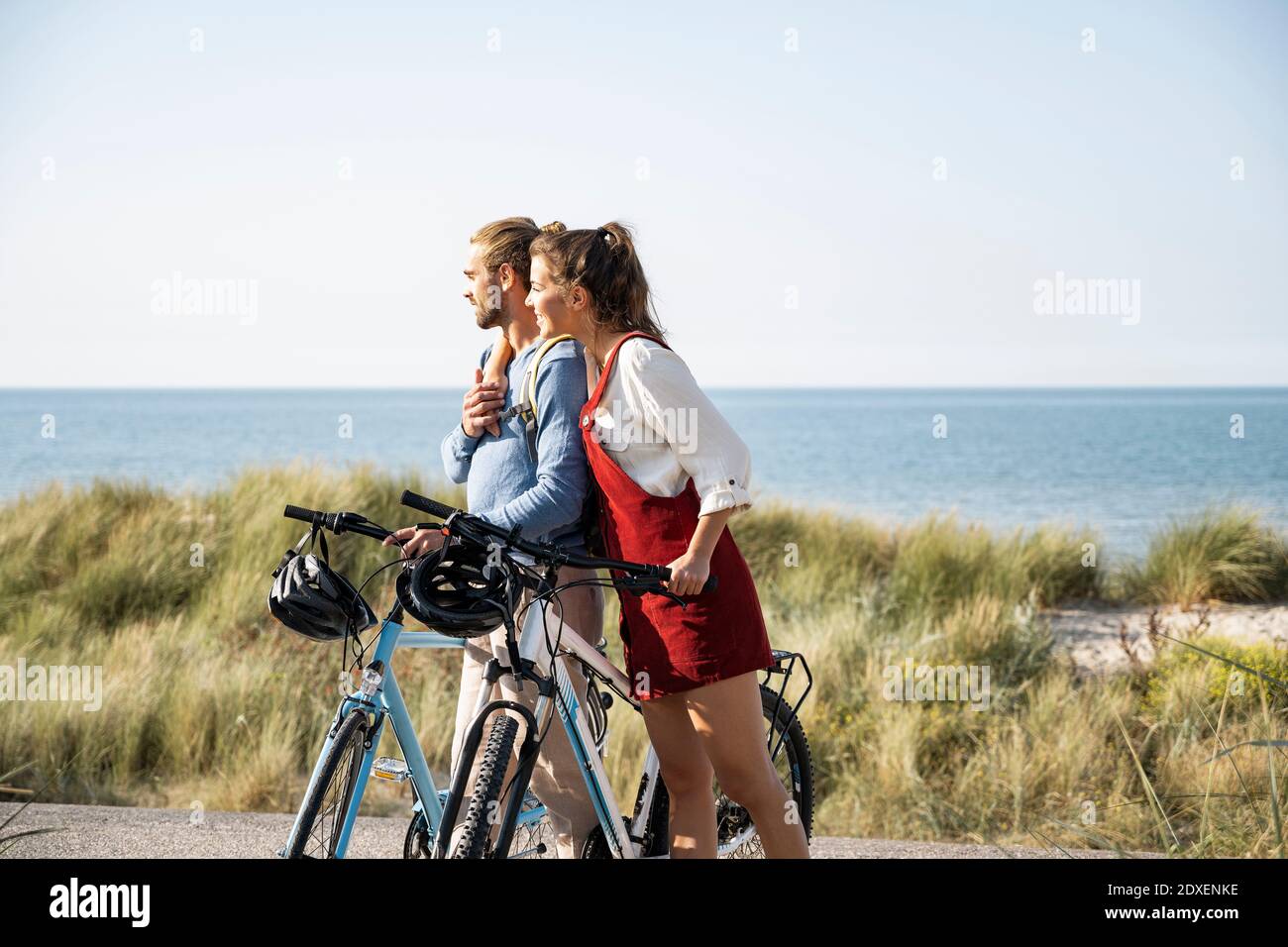 Fidanzata con braccio intorno al ragazzo ammirando la vista sul mare mentre si è in piedi con biciclette in spiaggia Foto Stock