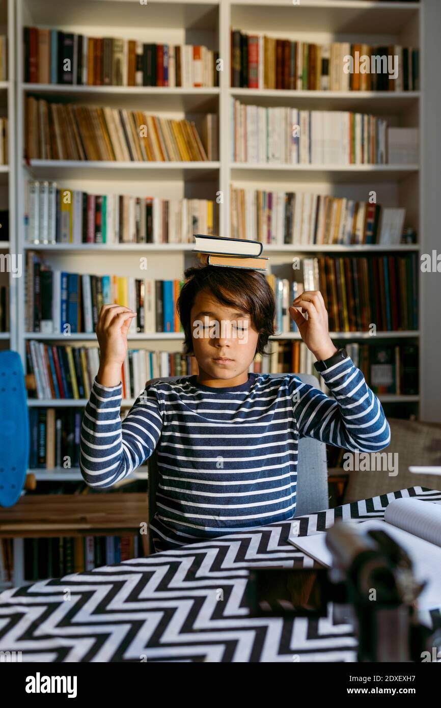 Ragazzo che equilibrano i libri sulla testa mentre vlogging video camera dentro soggiorno Foto Stock