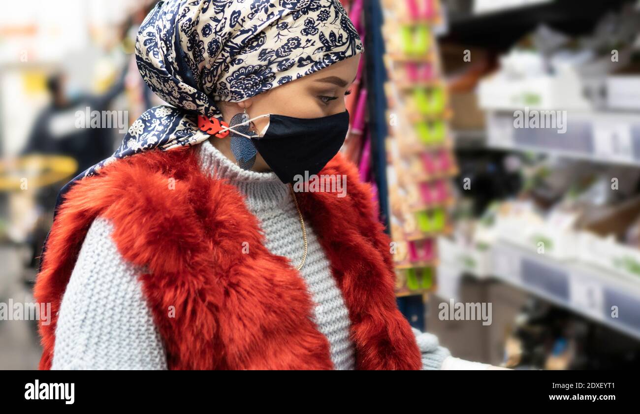 Giovane donna che indossa il copricapo e maschera protettiva shopping mentre in piedi al supermercato Foto Stock