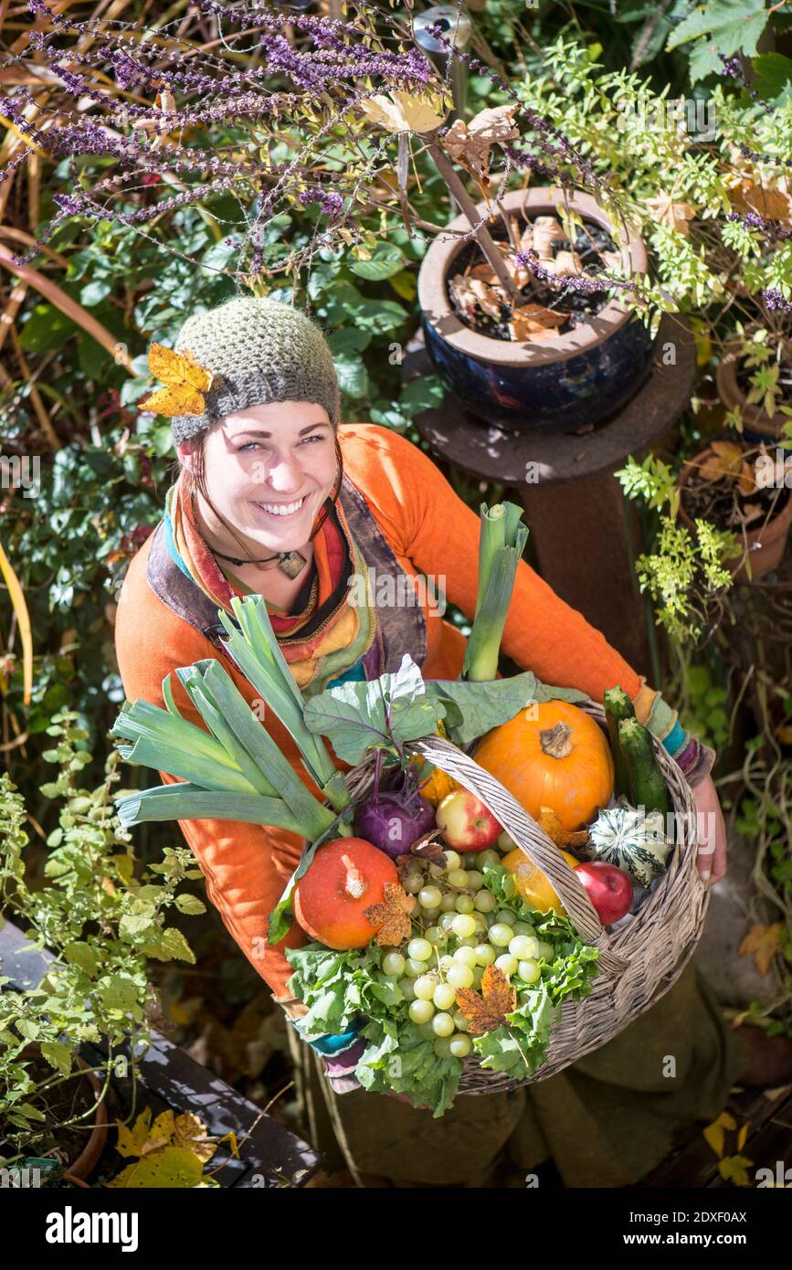 Donna sorridente che porta cesto di frutta e verdura mentre si trova in piedi giardino Foto Stock