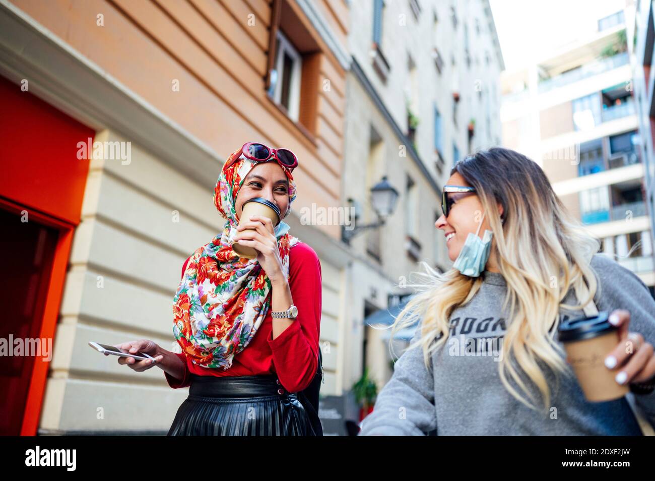 Sorridendo amici femminili con tazze usa e getta che camminano in città durante COVID-19 Foto Stock