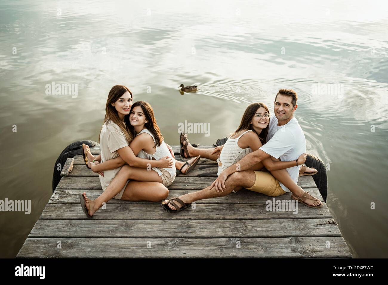Le figlie abbracciano madre e padre mentre si siedono sul molo contro lago Foto Stock