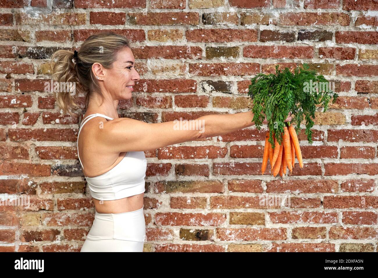 Donna matura sorridente che tiene mazzo di carote in cucina vicino muro di mattoni Foto Stock