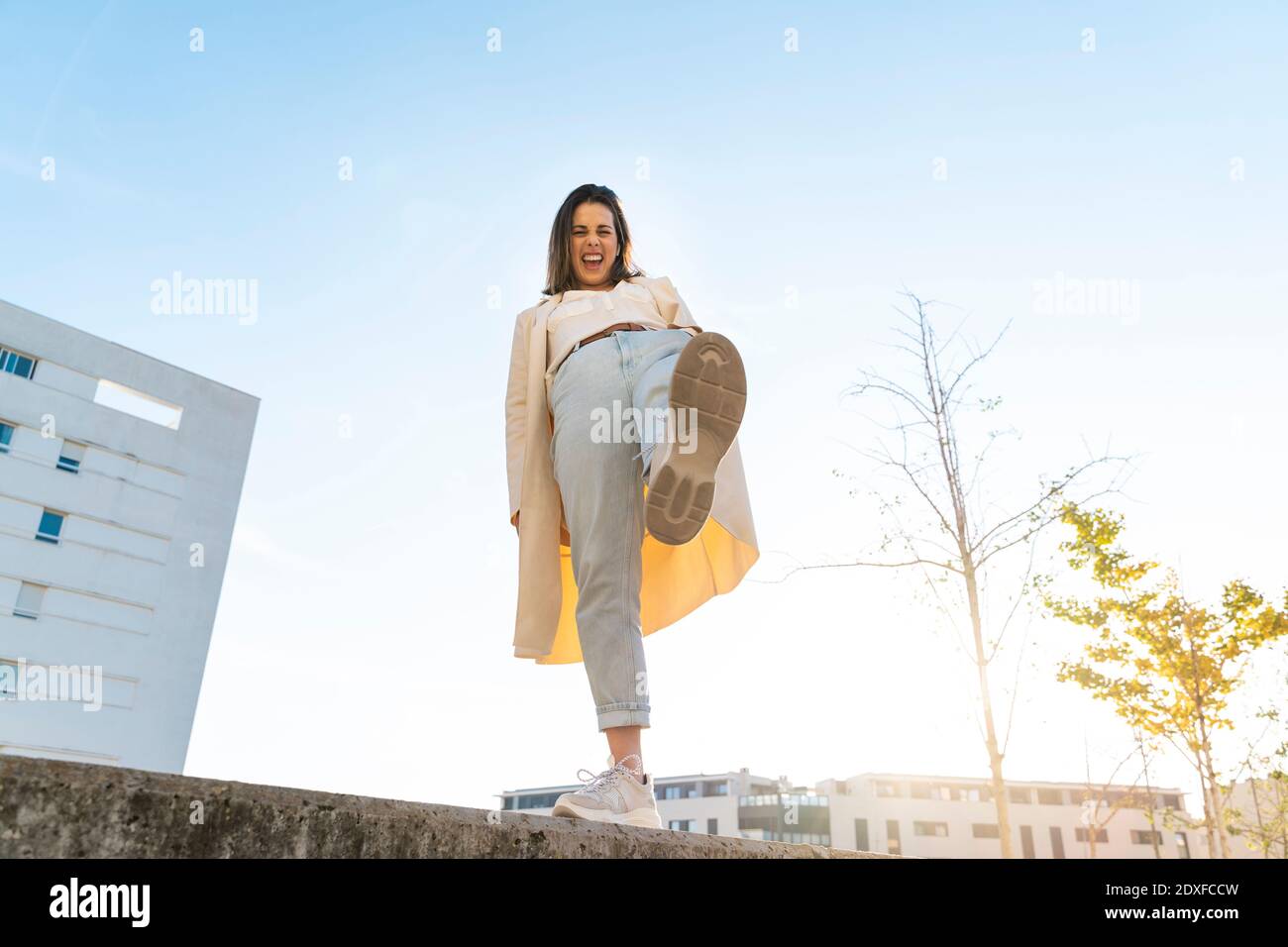 Donna d'affari allegra con gamba in piedi su muro di ritegno in città contro il cielo Foto Stock