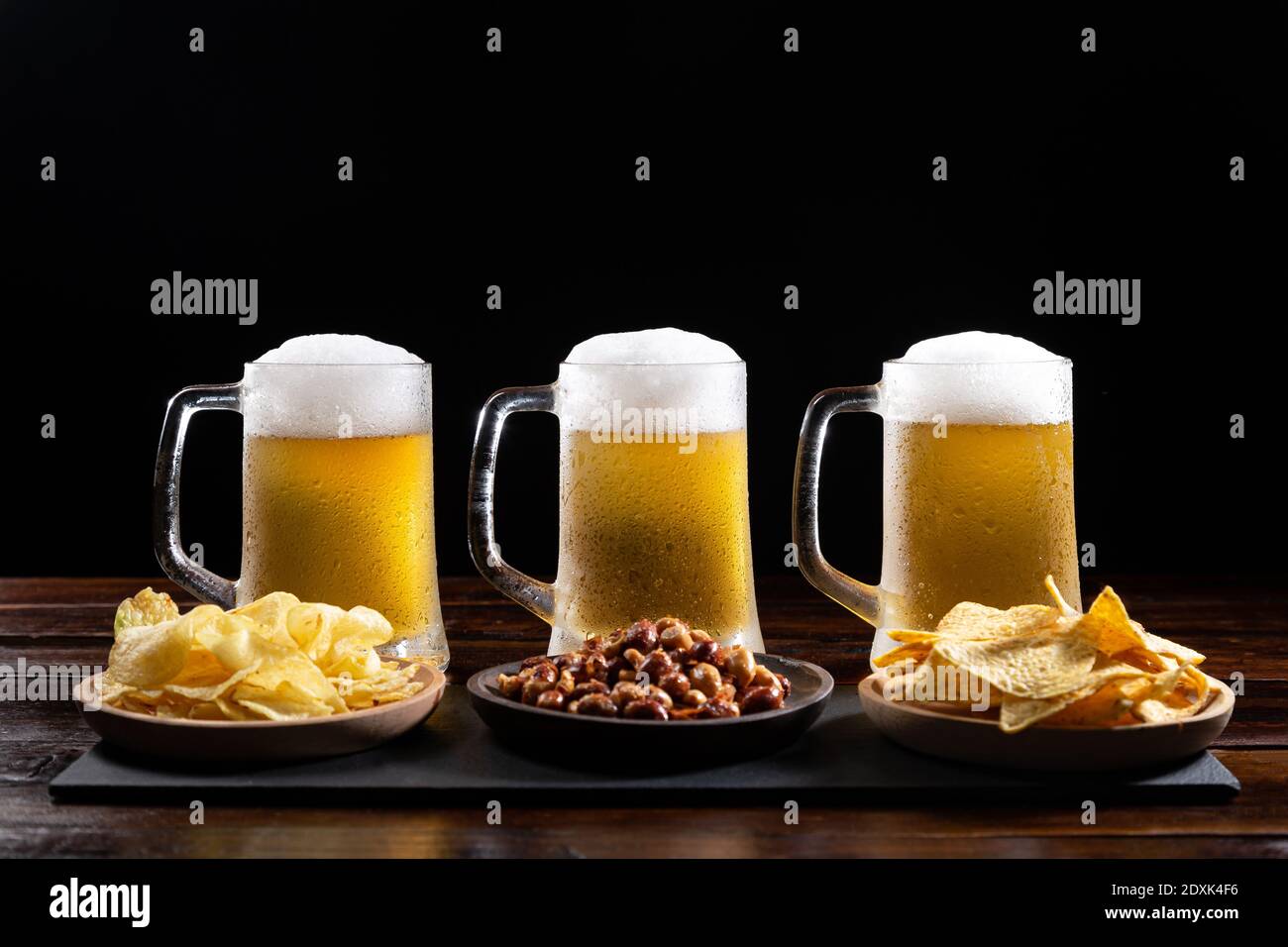 Tre tazze di birra ghiacciate e piatti con antipasti su base di legno su sfondo nero. Foto Stock