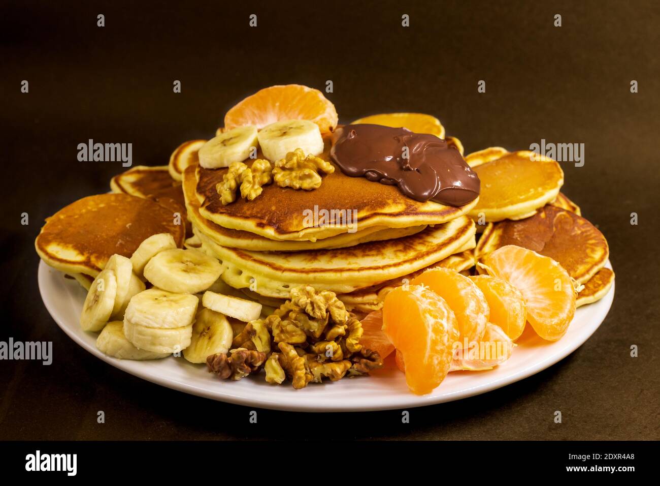 Piccole e grandi pancake ben cotti serviti da un piatto bianco con crema di nocciole al cioccolato, noce, mandarino e banana Foto Stock