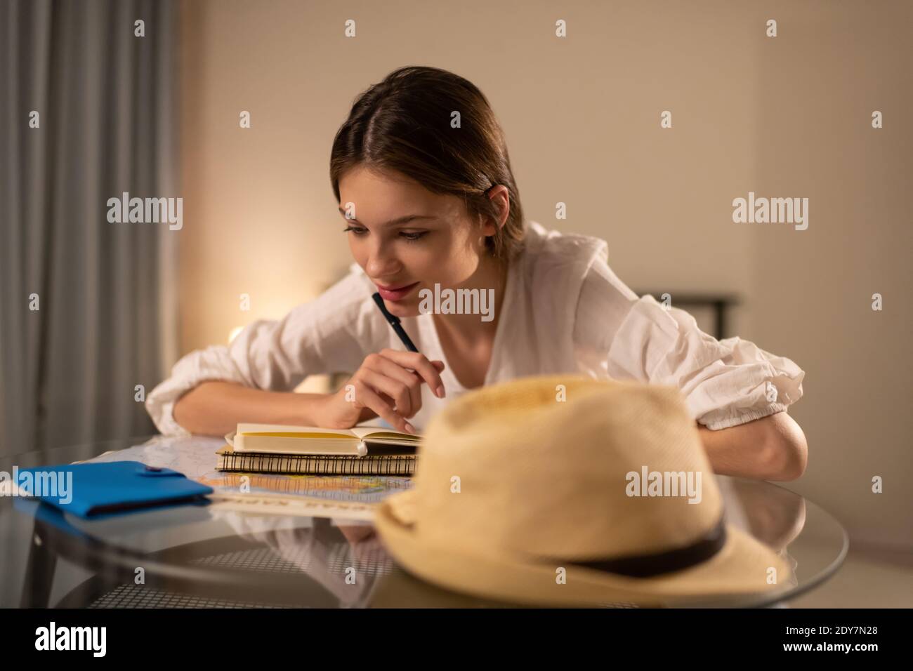 Ragazza felice con taccuino e penna che esamina la mappa e. pensare mentre si siede al tavolo a casa e pianificare un viaggio percorso Foto Stock