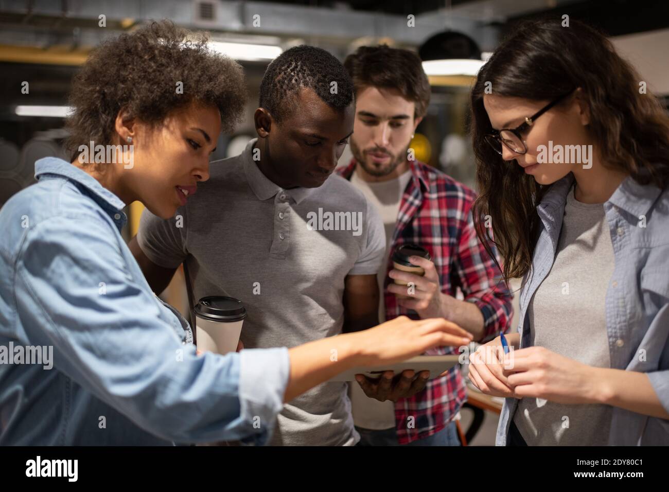 Gruppo di diversi colleghi in abiti casual e con tazze di bevande per andare a scorrere i dati sul tablet mentre si lavora su progetto in ufficio insieme Foto Stock