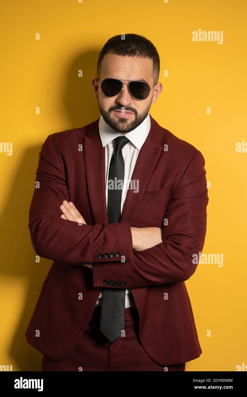 Uomo bello serio in colore bordeaux vestito e occhiali da sole cercando alla telecamera con le mani piegate isolate su sfondo giallo Foto Stock