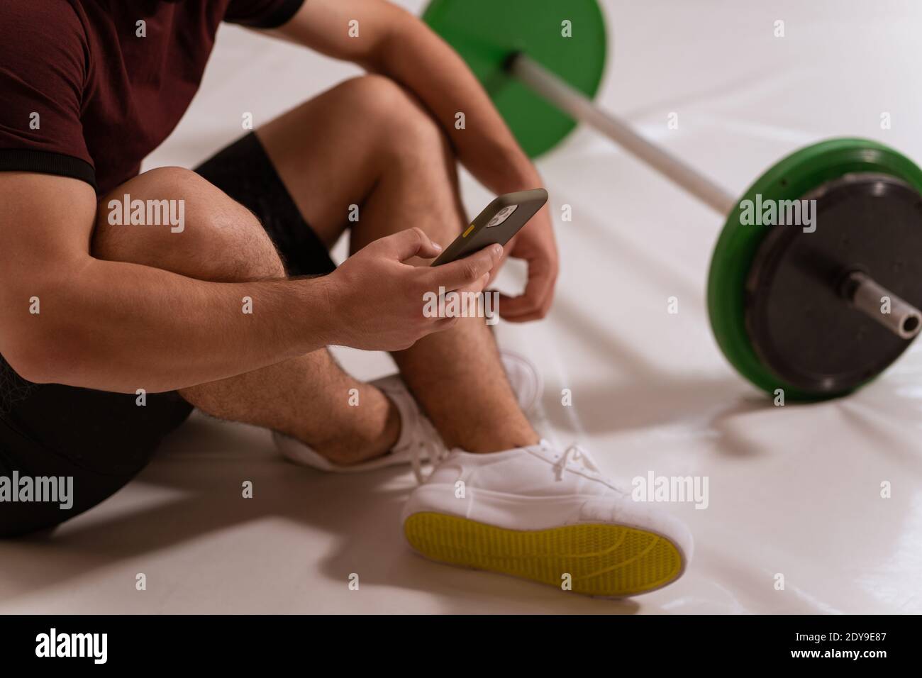 Body close up shot di un giovane uomo con smartphone in mano navigando online o testando seduto sul pavimento, tono nero e verde barbell fitness, attrezzature Foto Stock