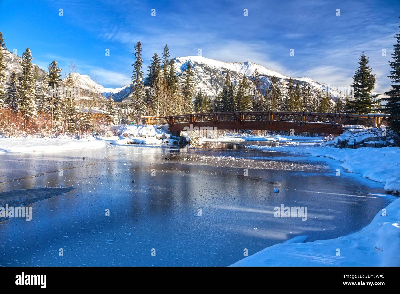 Ponte pedonale in legno sul ghiaccio del fiume ghiacciato, cime innevate delle Montagne Rocciose, skyline blu. Canadian Rockies Winter Landscape Banff National Park, Alberta Foto Stock
