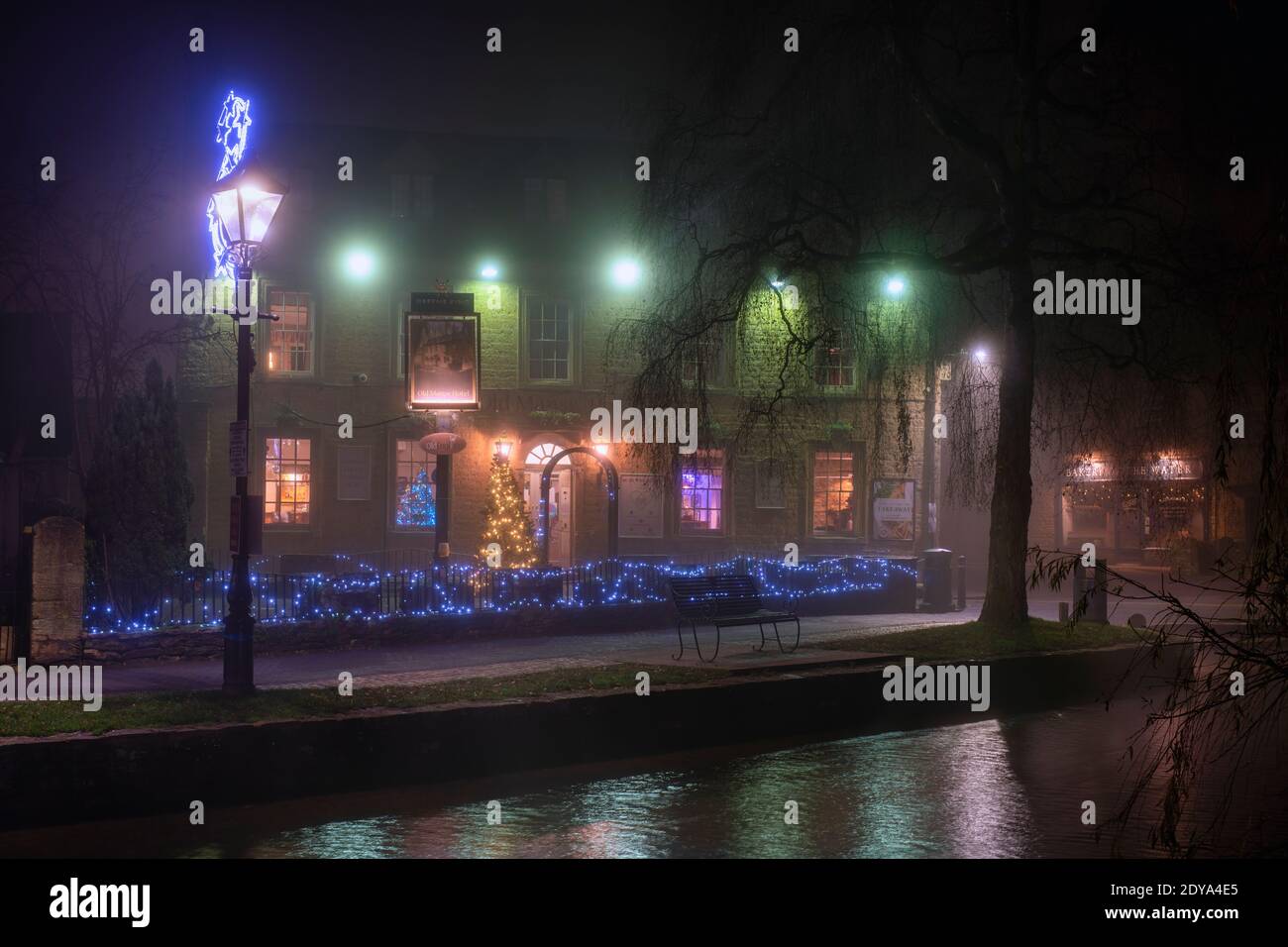 Albero di Natale e luci nella nebbia fuori dal vecchio hotel manse di notte. Bourton on the Water, Cotswolds, Gloucestershire, Inghilterra Foto Stock