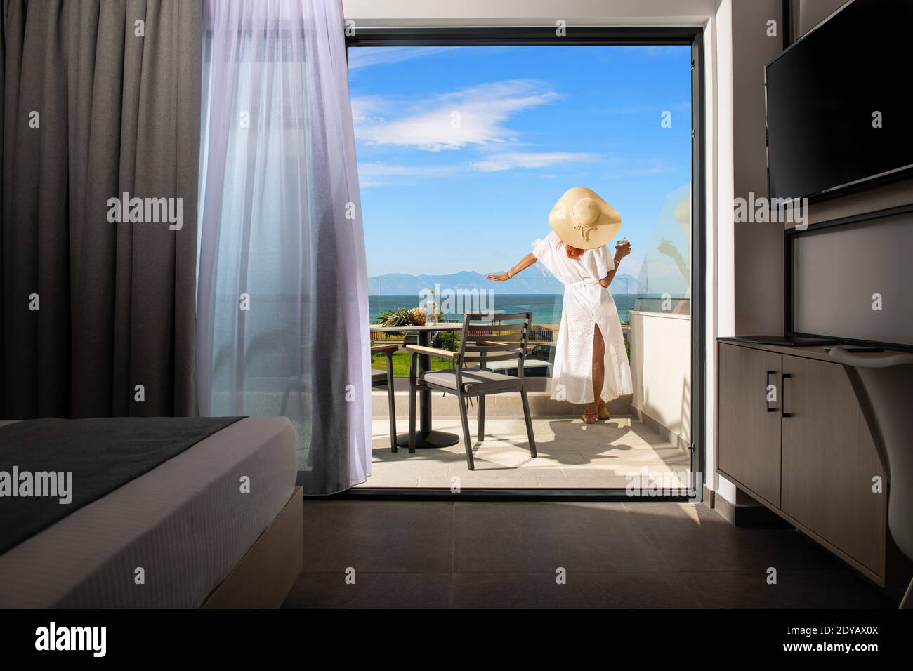 Vista posteriore di giovane donna in bianco abito estivo e. cappello di paglia sulla terrazza del balcone aperto della camera dell'hotel resort con vista sul mare Foto Stock