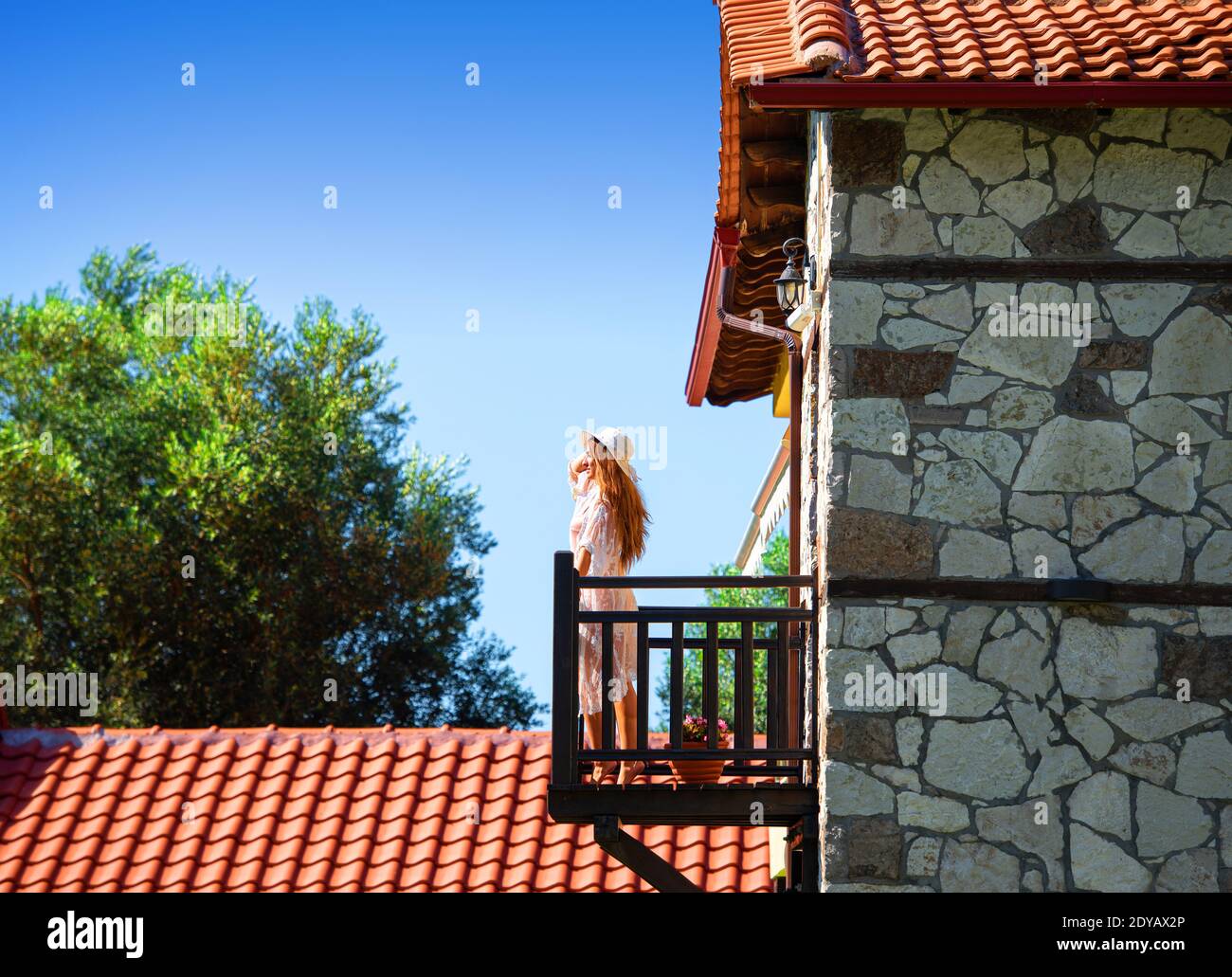 Vista laterale di giovane donna in piedi sul balcone di legno di vecchia casa di ciottoli con tetto di tegole rosse in estate paesaggio di campagna Foto Stock