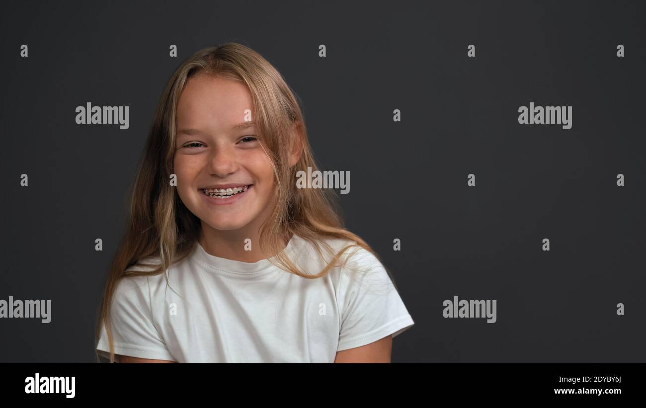 Felice ragazza sorridente con i capelli biondi in piedi guardando la fotocamera indossa una t-shirt bianca isolata su grigio scuro o. sfondo nero Foto Stock