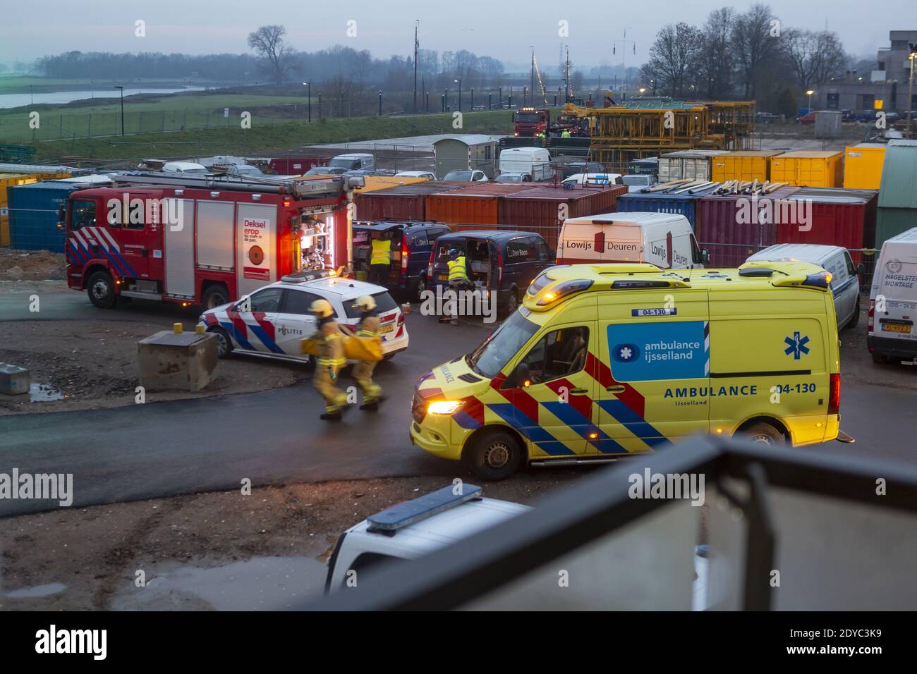Ambulanza in arrivo e vigili del fuoco che scorrono verso la scena di un Incidente in un cantiere con il fiume IJssel in lo sfondo all'alba Foto Stock