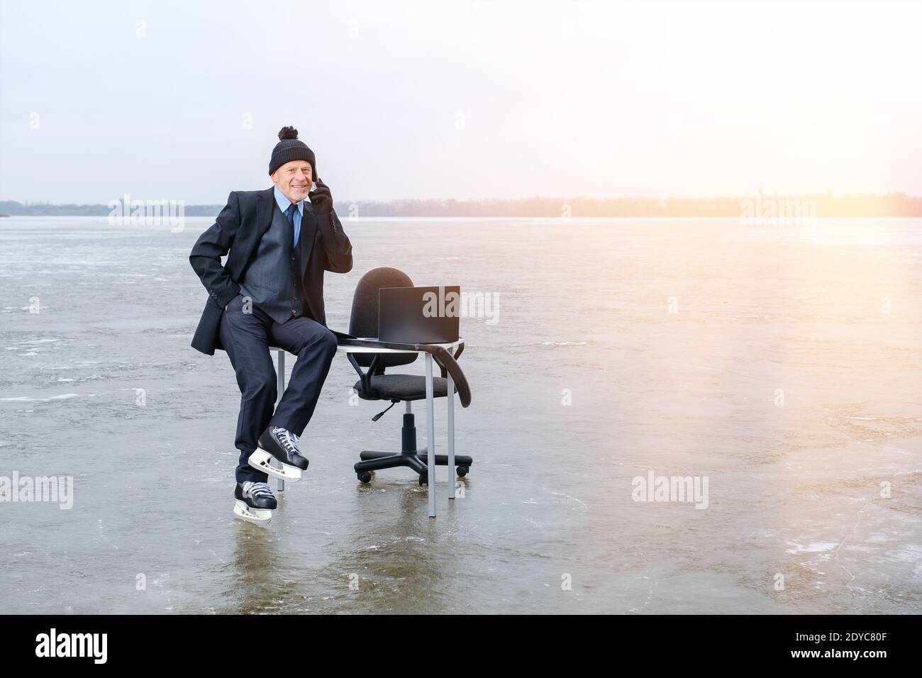 uomo d'affari sorridente in costume e pattini si siede sul tavolo sul lago ghiacciato Foto Stock