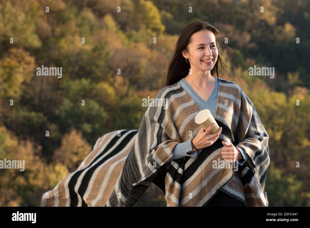 Bel moman in un poncho e un caffè in mano su sfondo natura. Bruna ragazza sorridente. Foto Stock