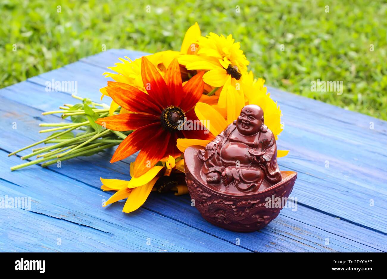Statuina di buddha in ceramica su tavole di legno blu invecchiate nel parco estivo con fiori di rudbeckia. Foto Stock