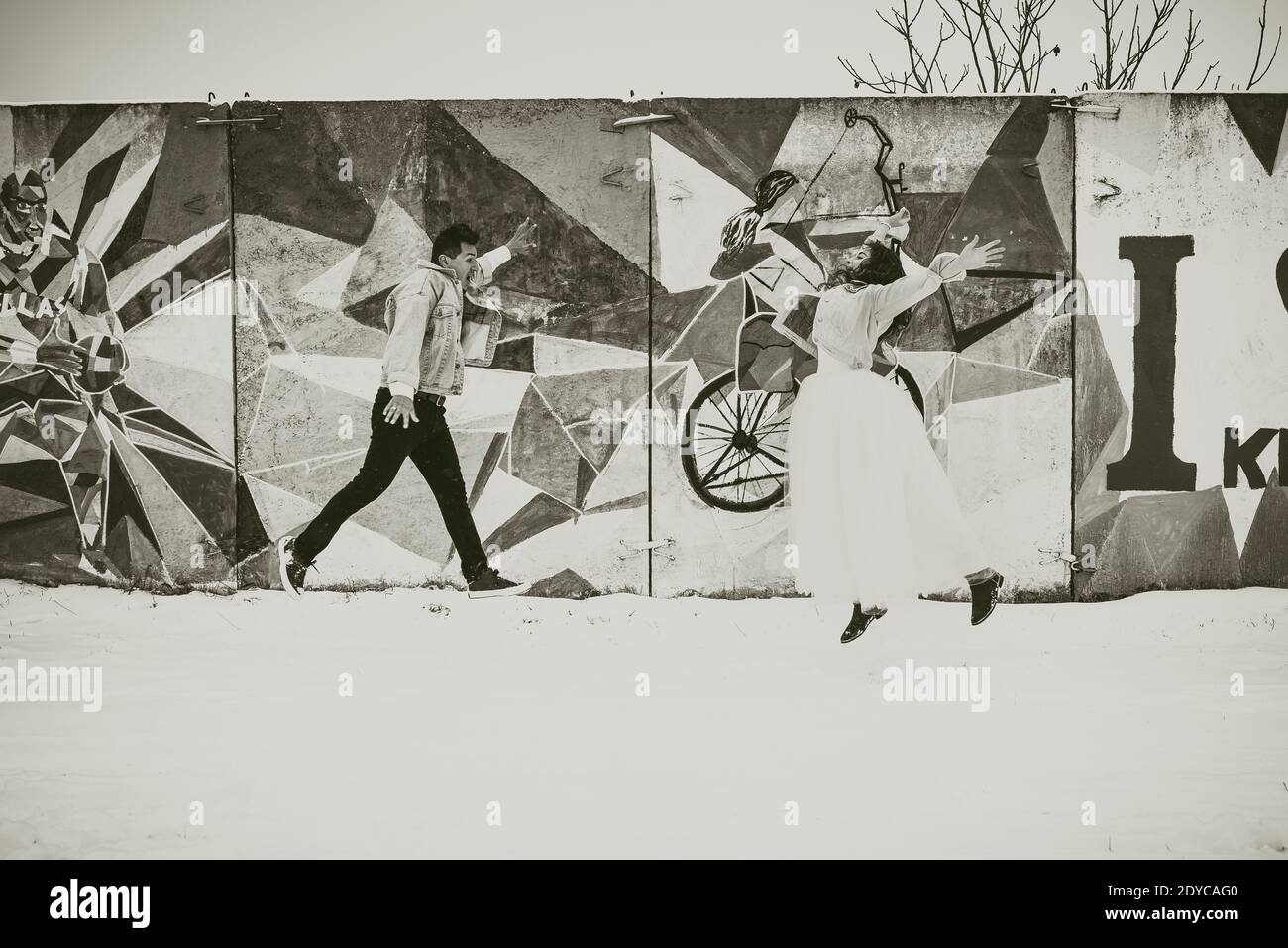 fresco giovane sposi in abiti da sposa e giacche in denim che saltano sullo sfondo del muro dipinto pop art street art in inverno nella neve. Foto Stock