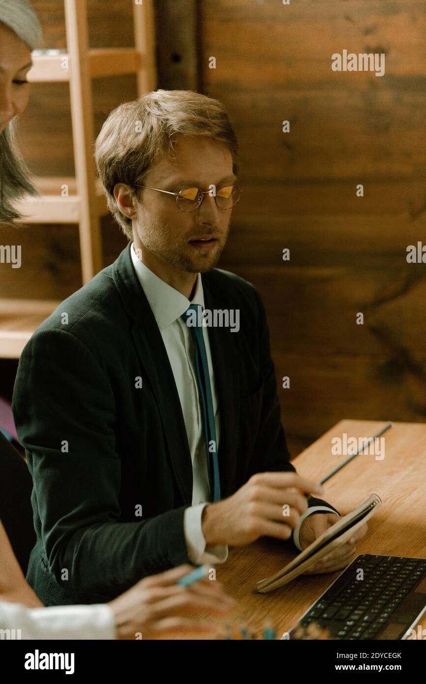 Giovane uomo con bicchieri freelance che lavora sul computer in ufficio. Uomo caucasico che mostra note ad un supervisore asiatico maturo. Operaio di collare bianco Foto Stock