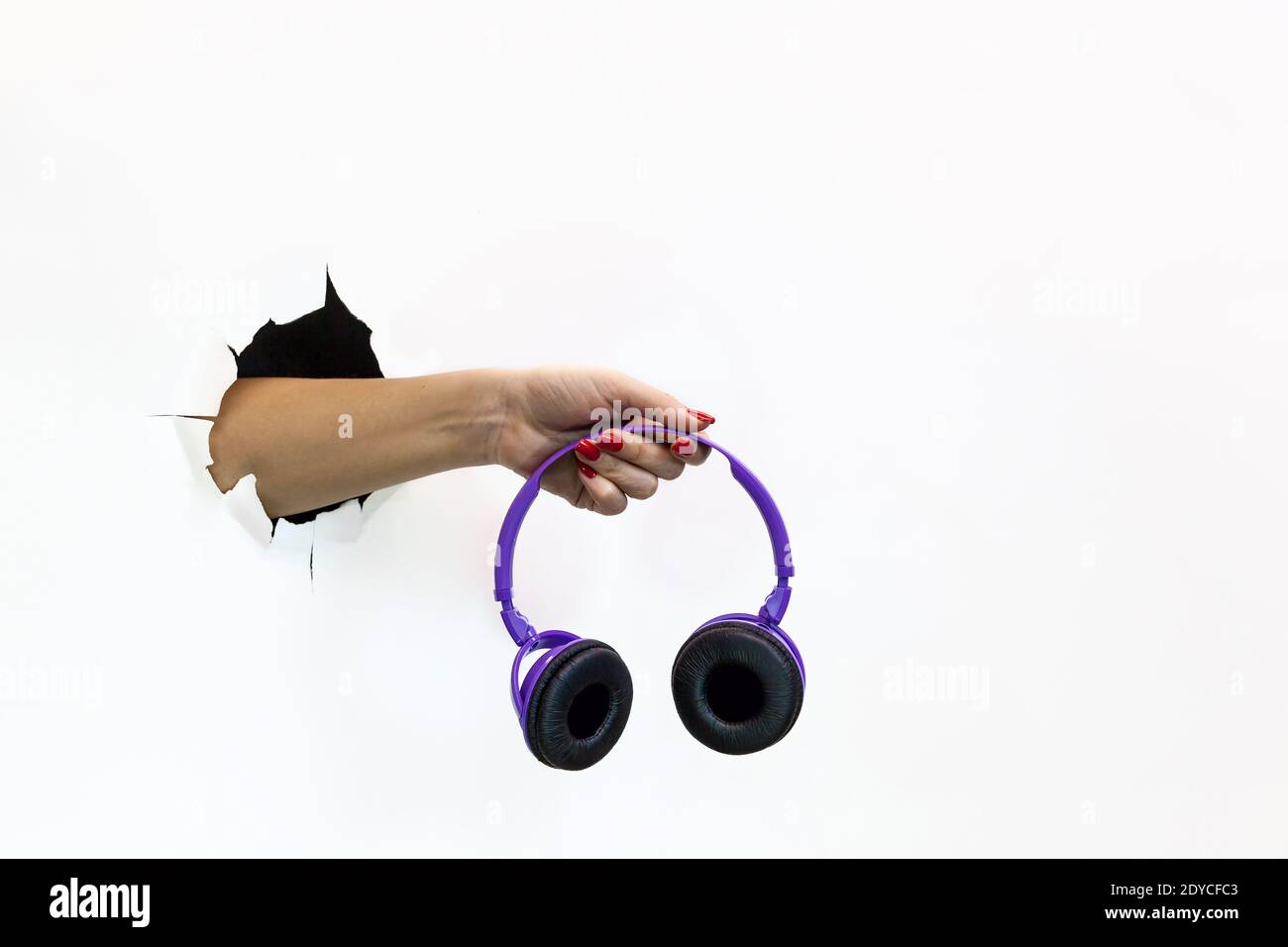 Una mano femminile con una manicure rossa su carta bianca strappata contiene una cuffia wireless viola. Passare la mano attraverso la carta bianca strappata Foto Stock