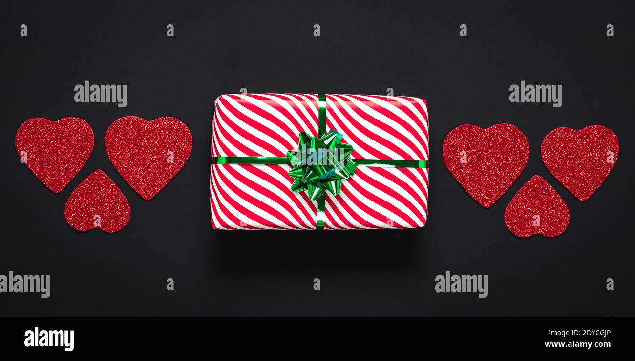 San Valentino, concetto d'amore. Romantica card panoramica scura con confezione regalo a righe bianche con arco verde. Regalo festivo e cuori rossi su una schiena nera Foto Stock